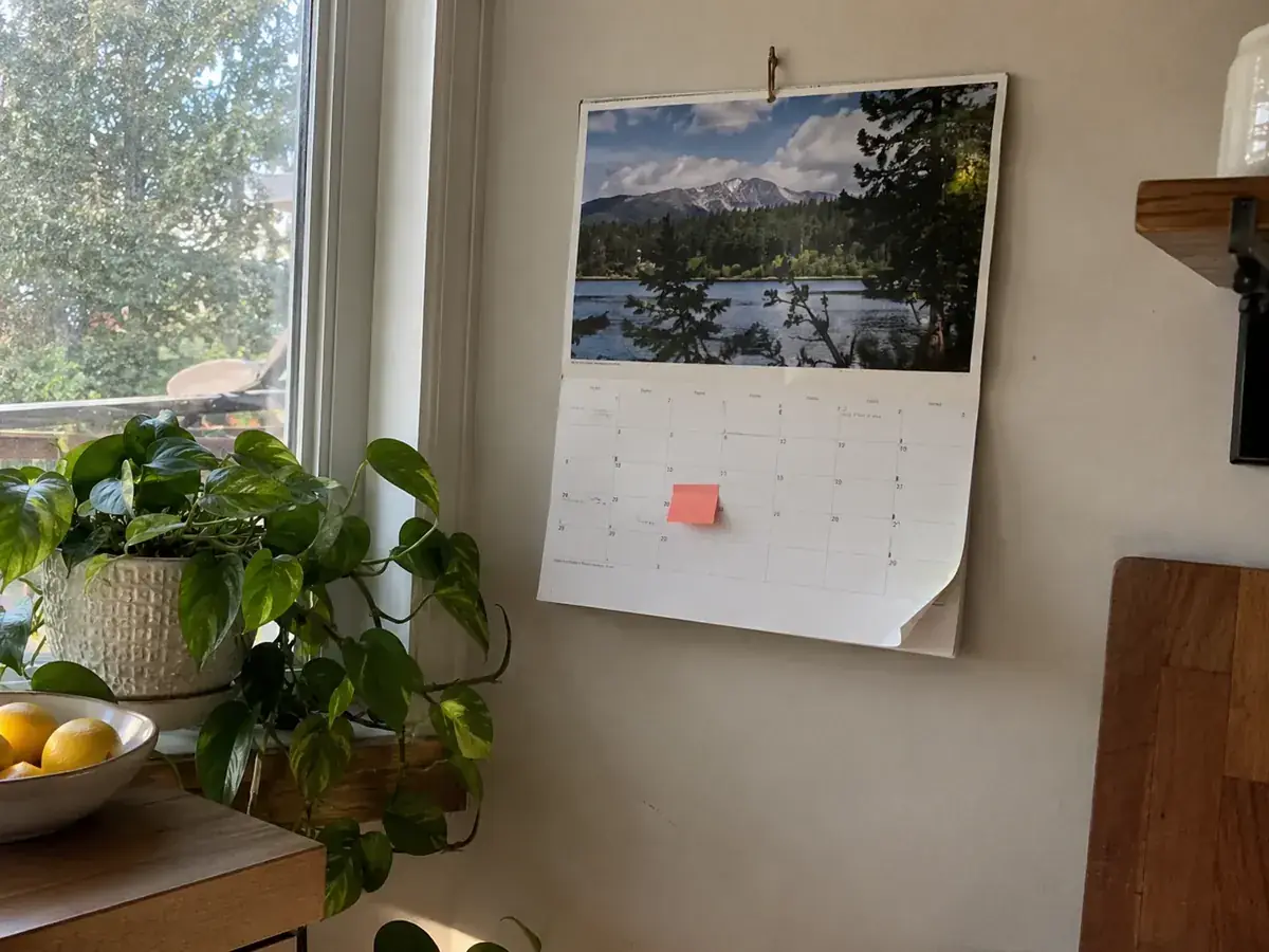 Imagen generada: Calendario de pared con una nota adhesiva junto a una ventana.