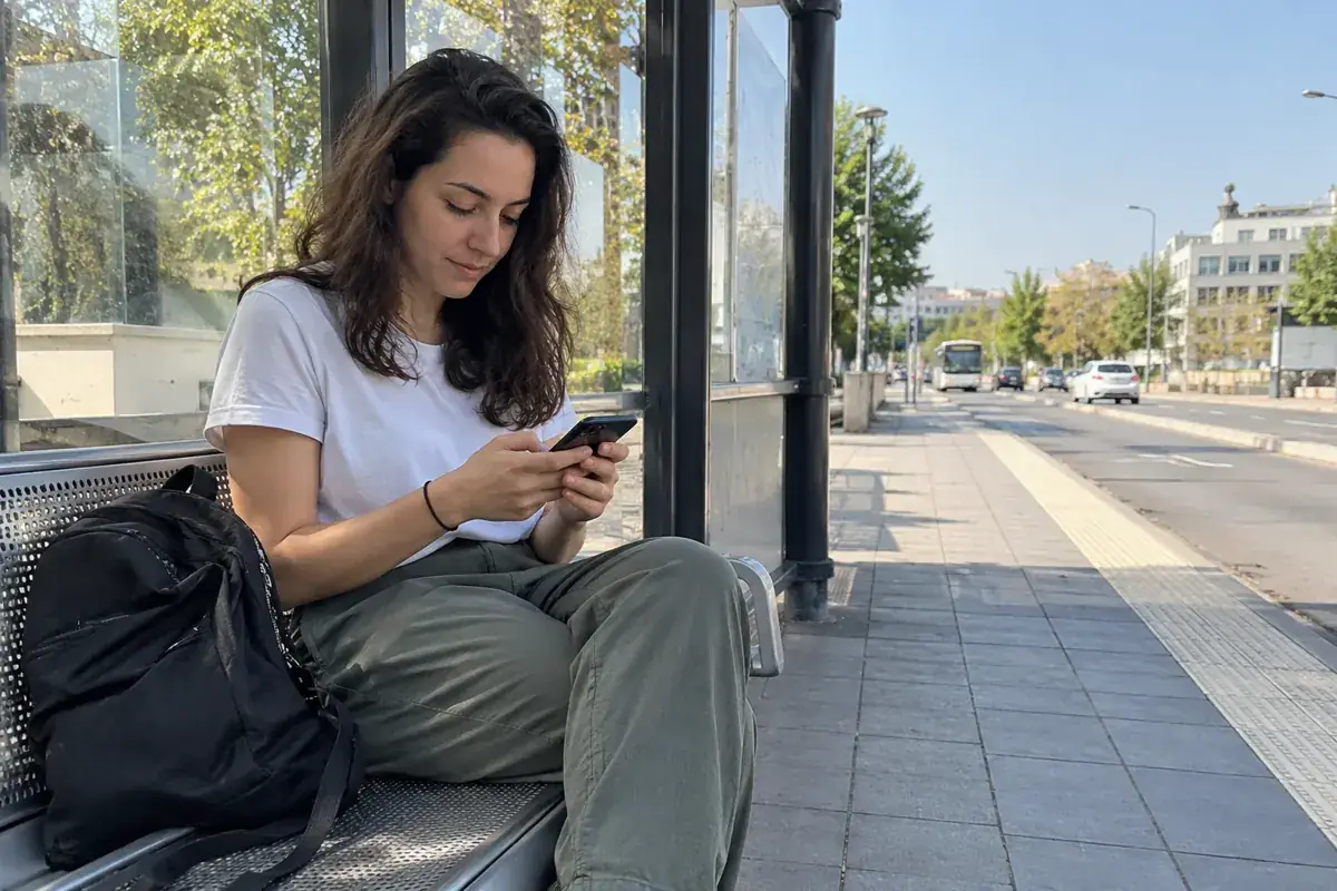 Imagen generada: Una mujer consulta su teléfono mientras espera en una parada de autobús.