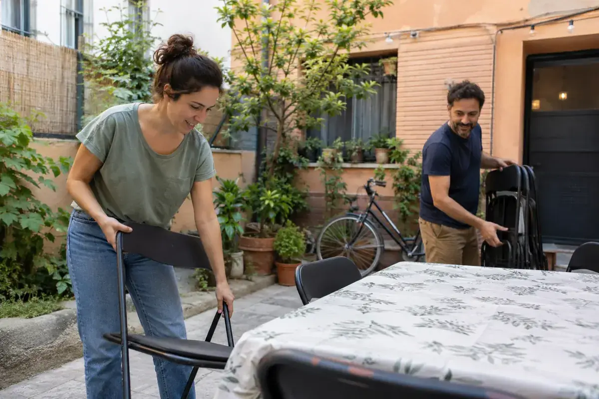 Imagen generada: Dos vecinos acomodan sillas plegables en un patio residencial.