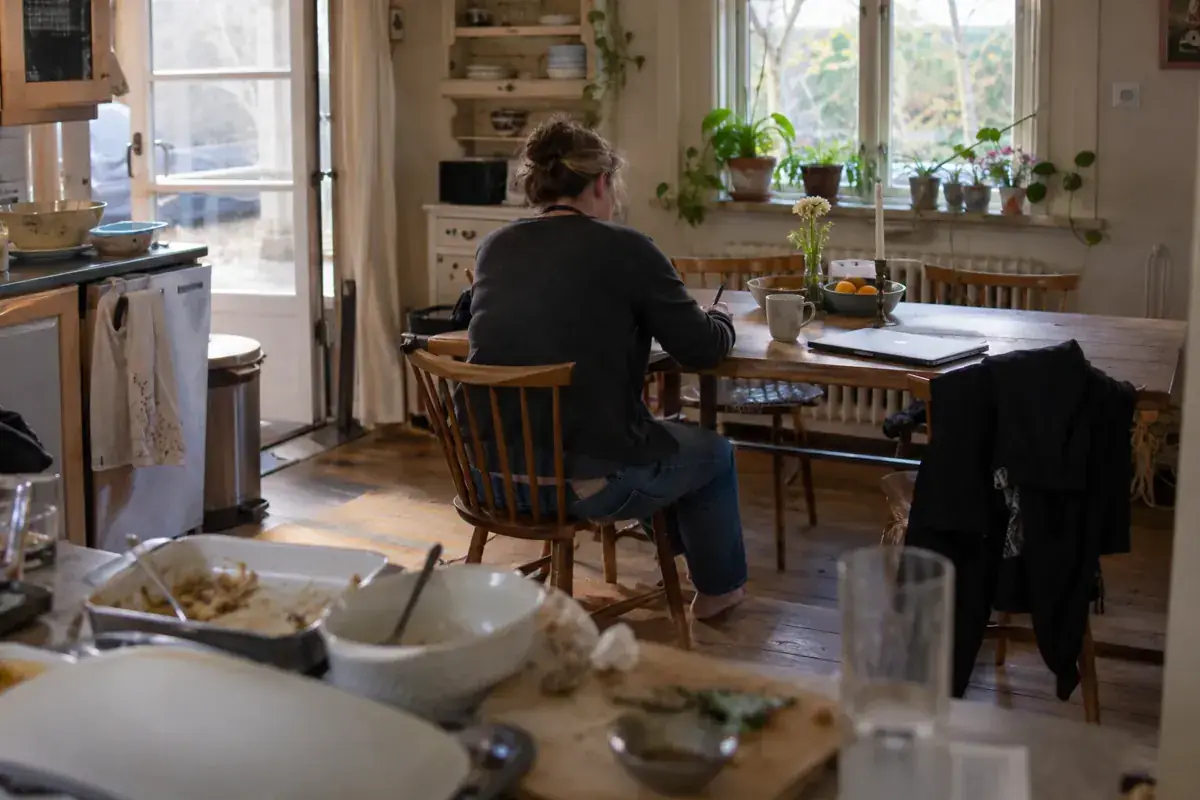 Persona de espaldas tomando notas en una mesa de cocina junto a una computadora y una taza.