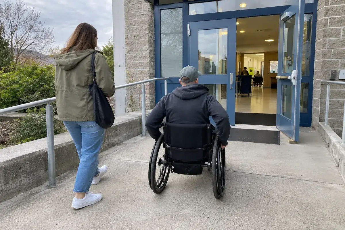 Una persona en silla de ruedas y otra a pie se acercan a una entrada con recorrido a nivel.