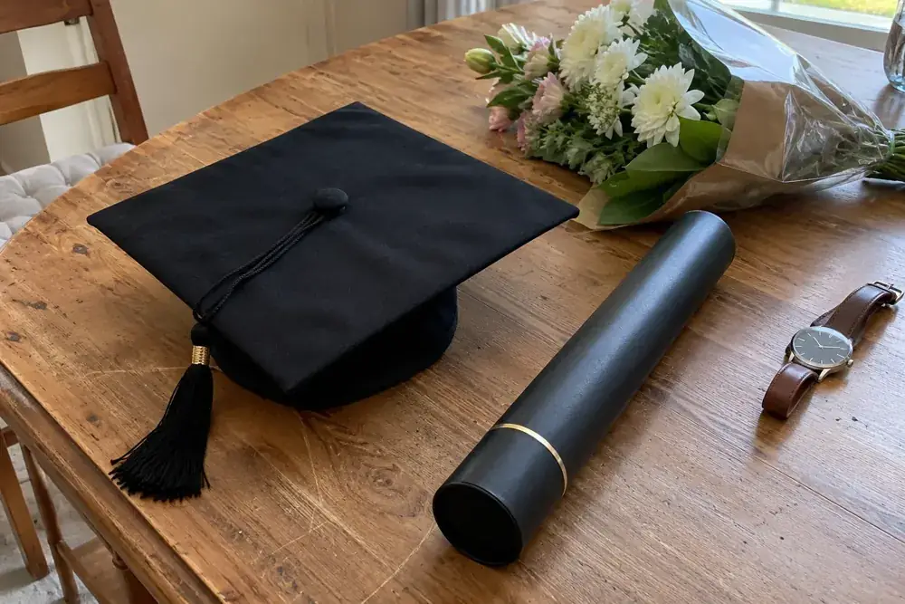 Birrete, tubo para diploma, reloj y flores sobre una mesa de madera.