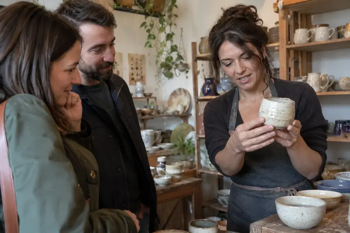 Imagen generada: Una artesana muestra una taza de cerámica a dos visitantes en su taller.