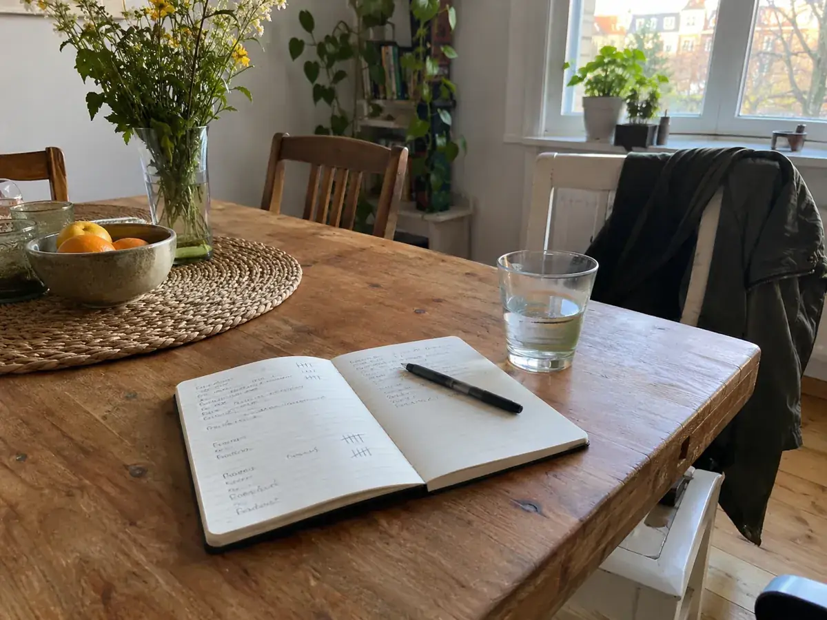 Imagen generada: Cuaderno abierto y bolígrafo sobre una mesa de comedor junto a un vaso de agua.