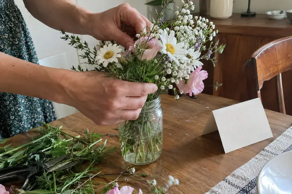 Imagen generada: Manos acomodando flores en un frasco de vidrio sobre una mesa de casa.