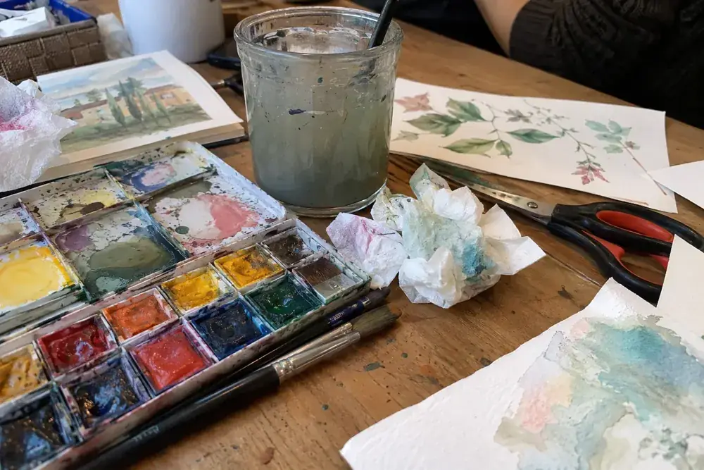 Imagen generada: Acuarelas usadas, vaso de agua y papeles sobre una mesa después de un taller.