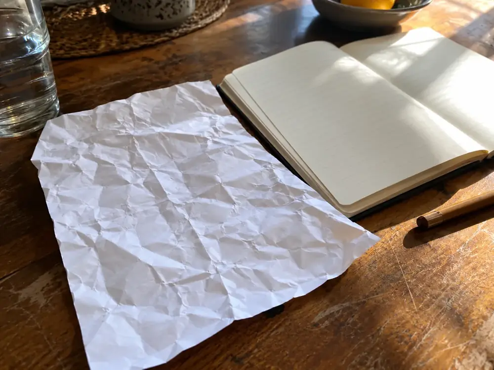 Imagen generada: Hoja arrugada sin texto, cuaderno abierto y un lápiz sobre una mesa.