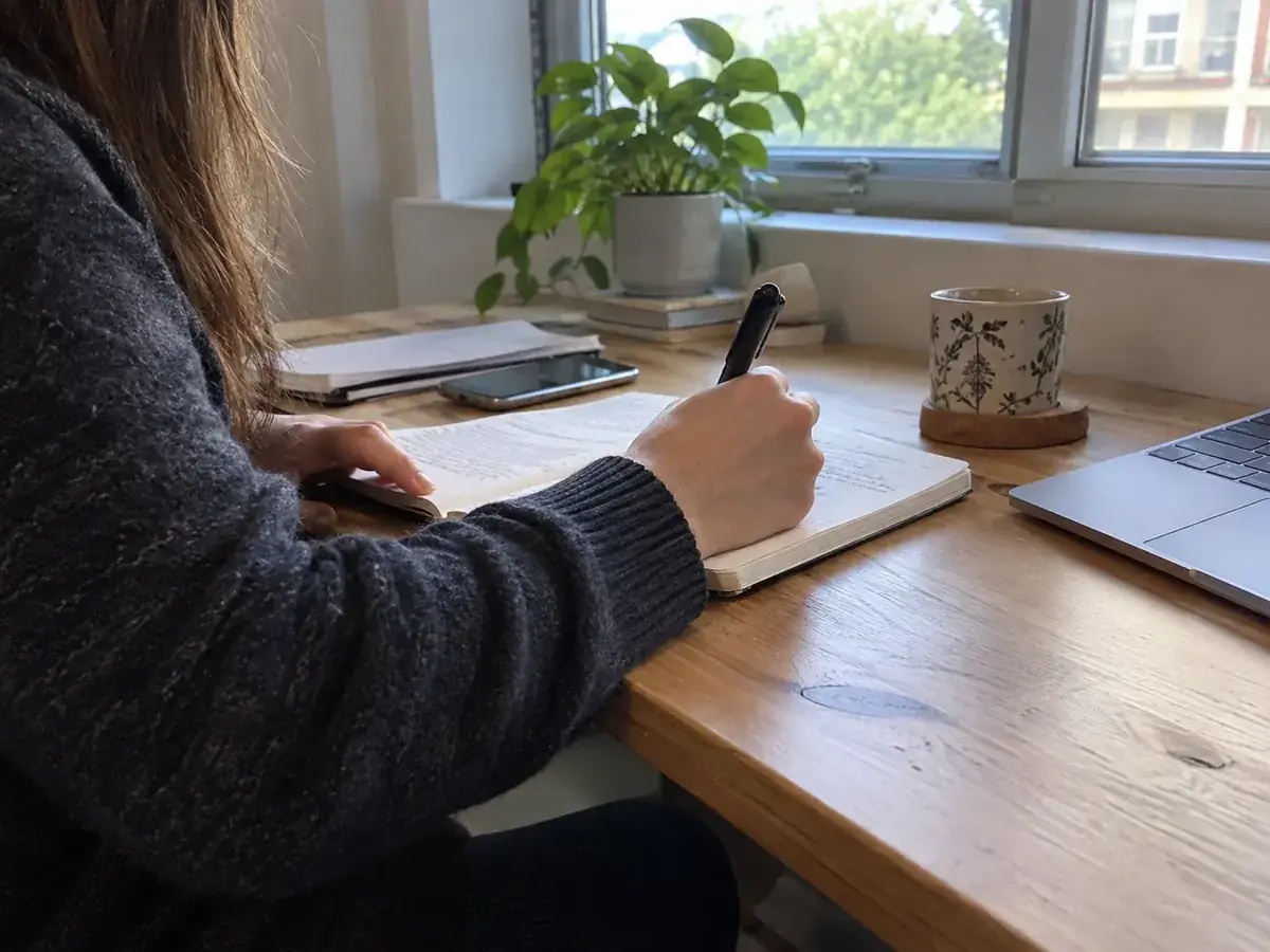 Imagen generada: Una persona escribe en un cuaderno junto a una computadora y una taza.